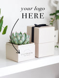 A styled product mockup showing a small potted succulent in a cream-colored square box with the text 'CLEARVIEW REAL ESTATE' printed on the front, alongside a larger matching gift box with a black ribbon. The image includes a black arrow and the text 'your logo HERE' indicating a customizable logo area. - Image 
