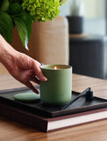Hand holding a matte sage green Lula's scented candle with a lit wick, lid, and black wick trimmer on a dark tray. - Image 