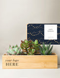 A wooden rectangular planter box containing three small succulent plants, with a navy blue gift sleeve behind it featuring gold string lights and a white 'Happy Holidays' label. The planter has placeholder text 'your logo HERE'. - Image 