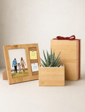 A wooden gift set featuring a photo frame with a family picture, a small succulent in a wooden planter, and a matching wooden gift box tied with a red ribbon, arranged on a light beige surface against a neutral background. - Image 