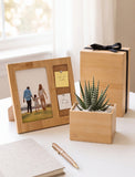 A styled tabletop scene with a light wood photo frame holding a family photo and a cork note holder, next to a small potted succulent in a wooden planter and a gift box tied with a black ribbon. A notebook and gold pen sit in the foreground on a white surface near a bright window. - Image 