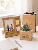 A styled wooden gift set on a white tabletop featuring a personalized photo frame, a small succulent in a branded wooden planter box, and a wooden gift box with a black ribbon bow. A notebook and gold pen are also present. - Image 