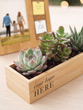 A rectangular light wood planter box containing several succulent plants, with the text 'your logo HERE' printed on the front. In the background, a wooden photo frame with a family picture and sticky notes sits on a light wood surface. - Image 