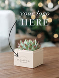 A small potted succulent sits in a cream-colored square box on a wooden table, with a blurred indoor background and overlaid text reading 'your logo HERE' plus a black arrow pointing to the box. The box is branded 'CLEARVIEW REAL ESTATE'. - Image 