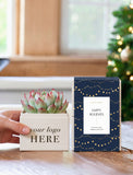 A hand holds a small potted succulent in a cream-colored square planter printed with 'your logo HERE' next to a navy blue gift box with gold string lights and a white 'Happy Holidays' label. The background shows a warm, softly blurred indoor setting with a Christmas tree. - Image 