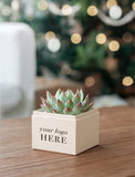 A small potted succulent in a cream-colored square planter box with the text 'your logo HERE' printed on the front, set on a wooden table with a blurred festive background. - Image 
