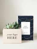 A holiday-themed gift box set featuring a cream-colored square box with 'your logo HERE' printed on the front, filled with succulents, next to a navy blue box with gold string lights and a white 'Happy Holidays' label. - Image 