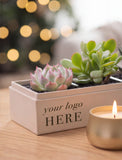 A rectangular beige gift box printed with 'your logo HERE' in black text, filled with two small succulent plants. The box sits on a wooden surface next to a lit gold candle, with a softly blurred holiday tree and warm lights in the background. - Image 