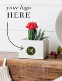 A white square planter box with a green circular logo sticker, holding a small red flower and succulent, displayed on a rustic wooden surface with a white paneled wall behind it. Text on the image reads 'your logo HERE' with an arrow pointing to the sticker area. - Image 