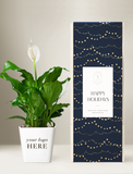 A potted peace lily plant in a white pot next to a tall rectangular gift box with a navy blue holiday design, gold string lights pattern, and a white label that reads 'Happy Holidays'. - Image 