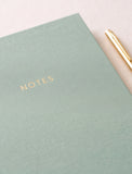 A close-up of a sage green textured notebook with gold foil lettering that reads 'NOTES', shown on a light beige surface with a gold pen partially visible at the top right. - Image 