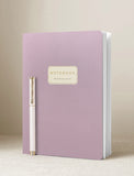 A lavender purple notebook with a gold-accented label on the front, standing upright on a light beige surface with a matching gold-and-white pen leaning against its left side. - Image 