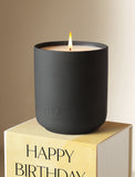 Lit Santal Noire candle in a matte black ceramic jar resting on a yellow Happy Birthday gift box. - Image 