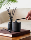 Matte black scented candle in a ceramic jar with a reed diffuser and wick trimmer on a brown coffee table book. - Image 
