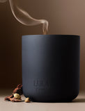 Matte black Lula's Candles jar with wisps of smoke, accented by cardamom pods and wood bark on a neutral background. - Image 