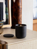 Lit Santal Noire scented candle in a matte black ceramic jar with a matching lid, sitting on a textured beige surface. - Image 