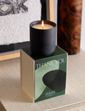 Lit Santal Noire candle in a matte black jar resting on a green Thank You gift box with abstract shapes. - Image 