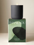 A lit matte black Lula's Santal Noire candle resting on a green Thank You gift box with abstract shapes. - Image 