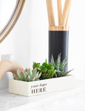 A rectangular cream-colored planter box with the text 'your logo HERE' in black, filled with various green succulents and small cacti, styled on a white marble surface next to a black reed diffuser with a copper-colored rim. - Image 