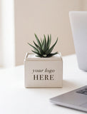 A small potted succulent in a square cream-colored planter box printed with the placeholder text 'your logo HERE', styled on a white desk next to a laptop. - Image 